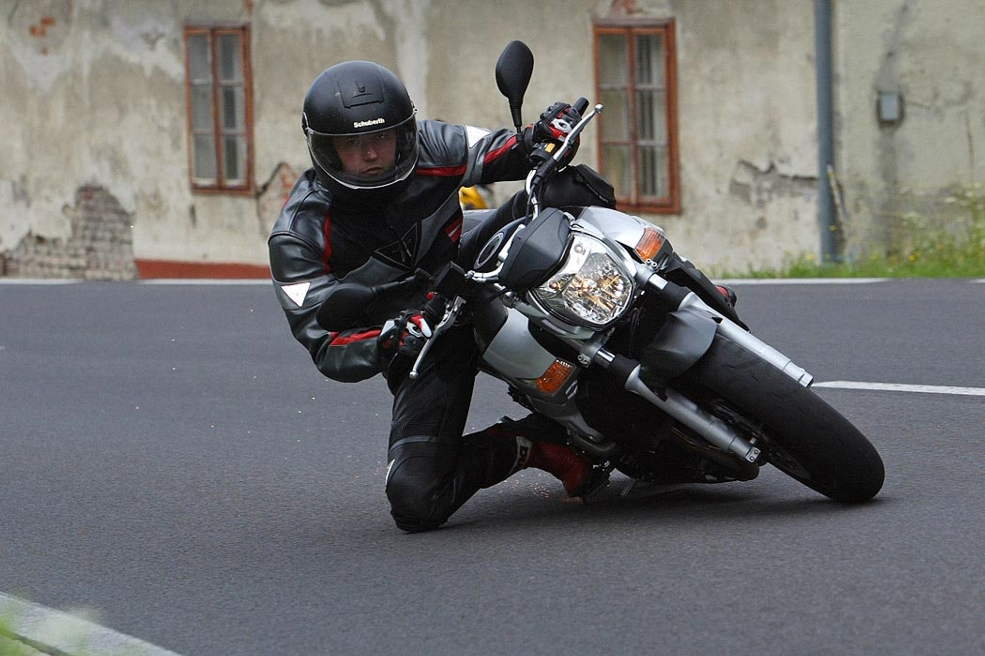NastyNils riding a Suzuki GSR600 through a spirited corner on asphalt, leaning the naked bike aggressively into the turn. Rider wears black leather jacket with red and white accents, full-face helmet. Weathered urban building visible soft in the background. Daylight, dry conditions. Action shot capturing the GSR600's cornering ability and rider technique.