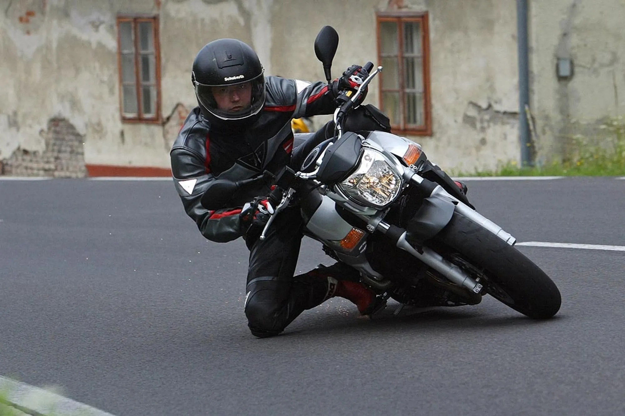 NastyNils riding a Suzuki GSR600 through a spirited corner on asphalt, leaning the naked bike aggressively into the turn. Rider wears black leather jacket with red and white accents, full-face helmet. Weathered urban building visible soft in the background. Daylight, dry conditions. Action shot capturing the GSR600's cornering ability and rider technique.