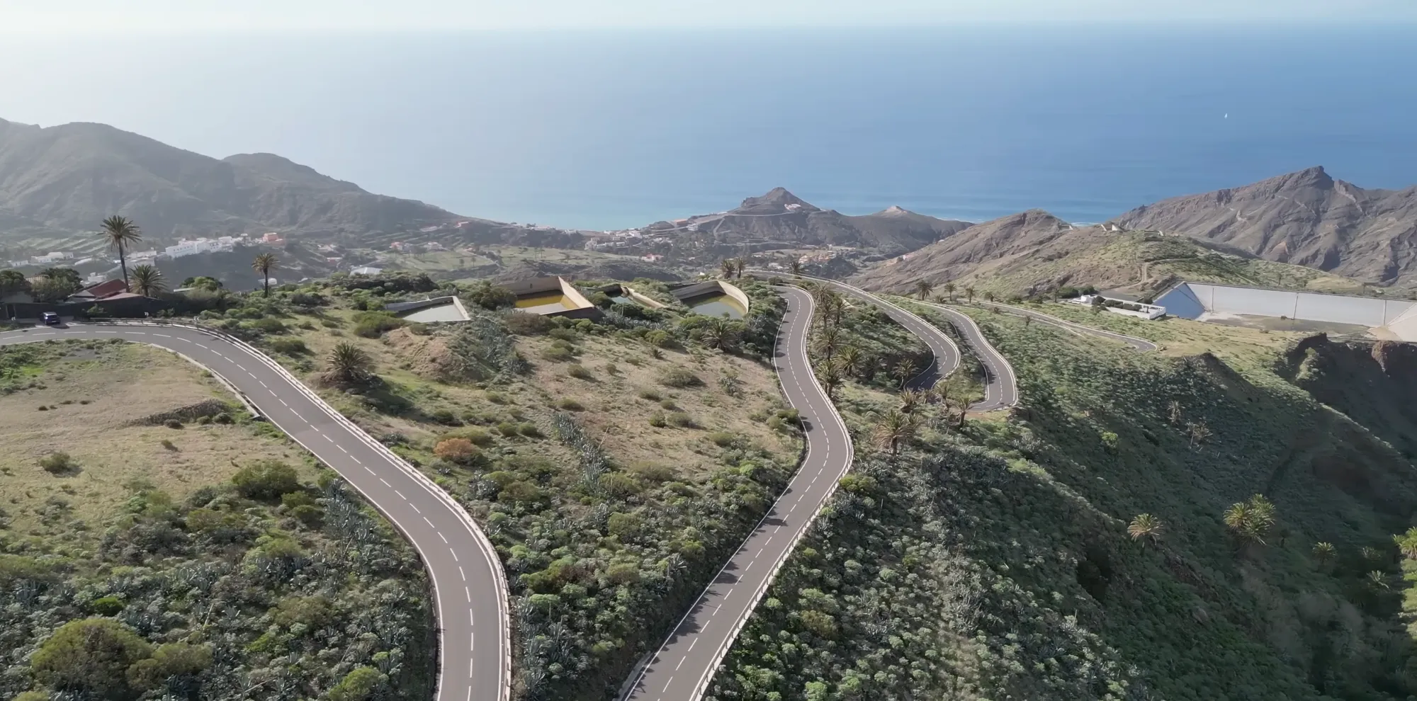 Aerial view of a winding asphalt road cutting through volcanic terrain on La Gomera, Canary Islands. The road curves through sparse green vegetation with rocky volcanic peaks visible in the background and a settled valley to the left. Clear lane markings, dry climate, partly cloudy sky.