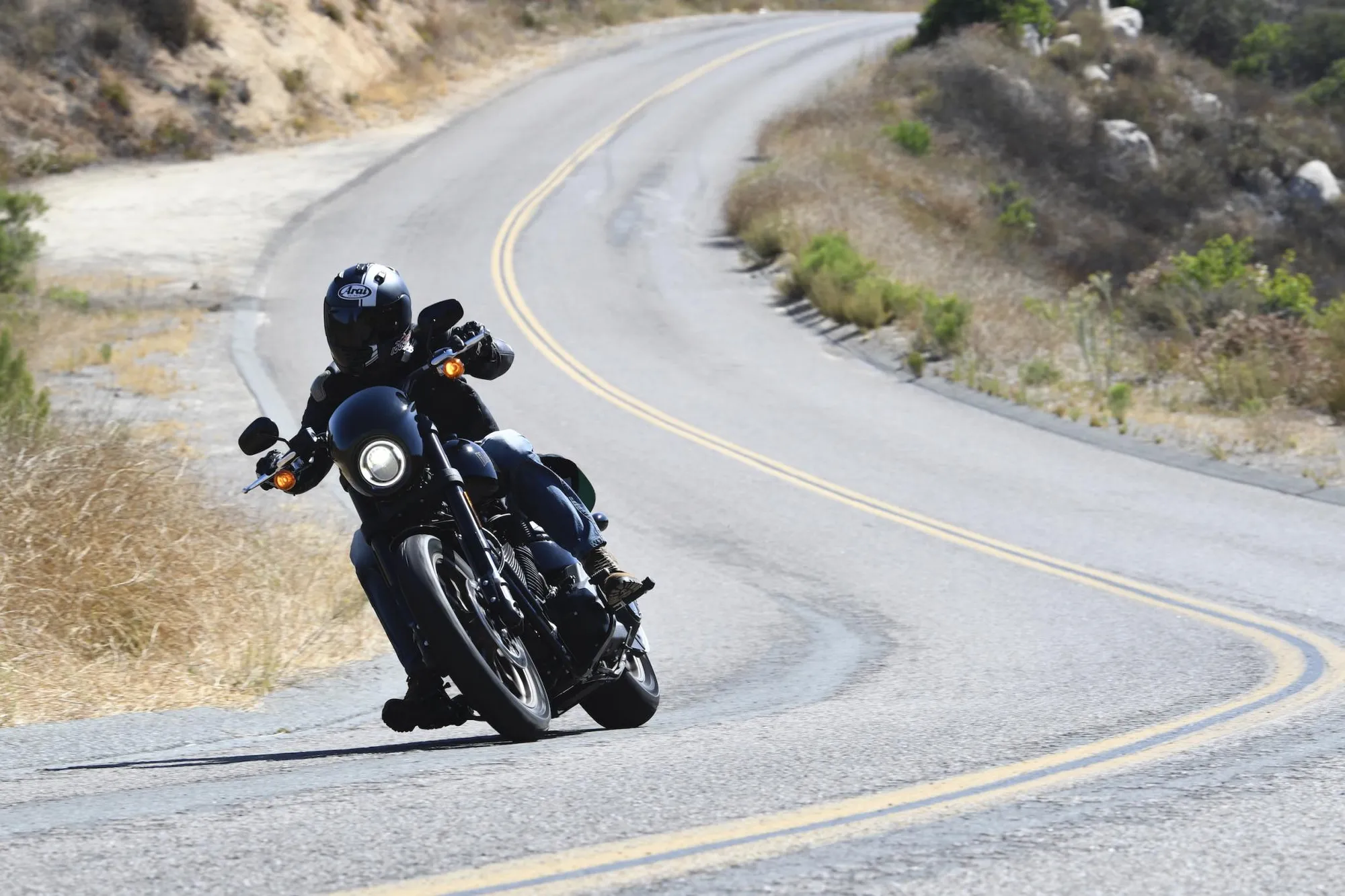 NastyNils riding a Harley-Davidson Low Rider S through a winding mountain road in California, leaning into a right-hand curve. Dry hills with sparse vegetation visible in the background. Daylight, clear road conditions, NastyNils in black full-face helmet and black leathers. Yellow center-line markings emphasize the curve ahead.