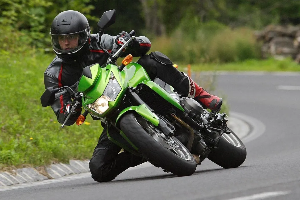 NastyNils riding a Kawasaki Z750 through a tight left-hand corner, knee touching the tarmac in a full kneedown. Bike finished in Kawasaki's signature lime green. Rider in black and red leather suit, black full-face helmet, and red Daytona boots. Green deciduous trees and roadside vegetation in the background suggest a European country road. Overcast daylight, dry tarmac, low-mounted camera angle.