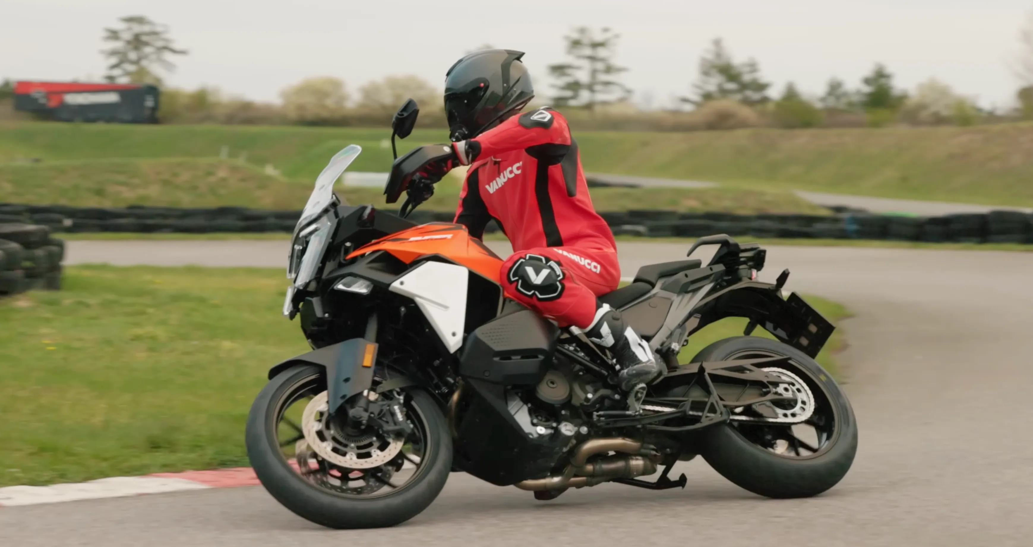 NastyNils rides the KTM 1390 Super Adventure S EVO (MY2025) through a tight paved corner on a supermoto circuit in Austria, leaned over in red Vanucci leathers, tire barriers and green track verges behind him — a big adventure bike pushed at sporting pace on asphalt.