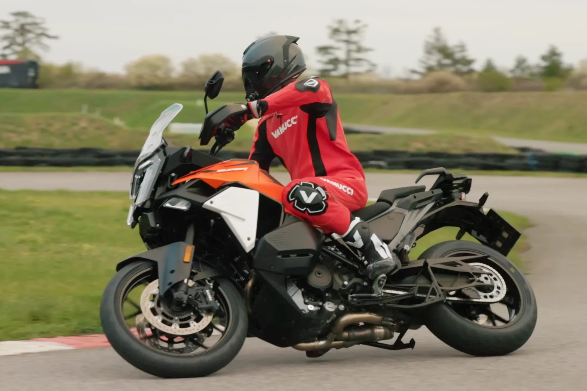 NastyNils rides the KTM 1390 Super Adventure S EVO (MY2025) through a tight paved corner on a supermoto circuit in Austria, leaned over in red Vanucci leathers, tire barriers and green track verges behind him — a big adventure bike pushed at sporting pace on asphalt.
