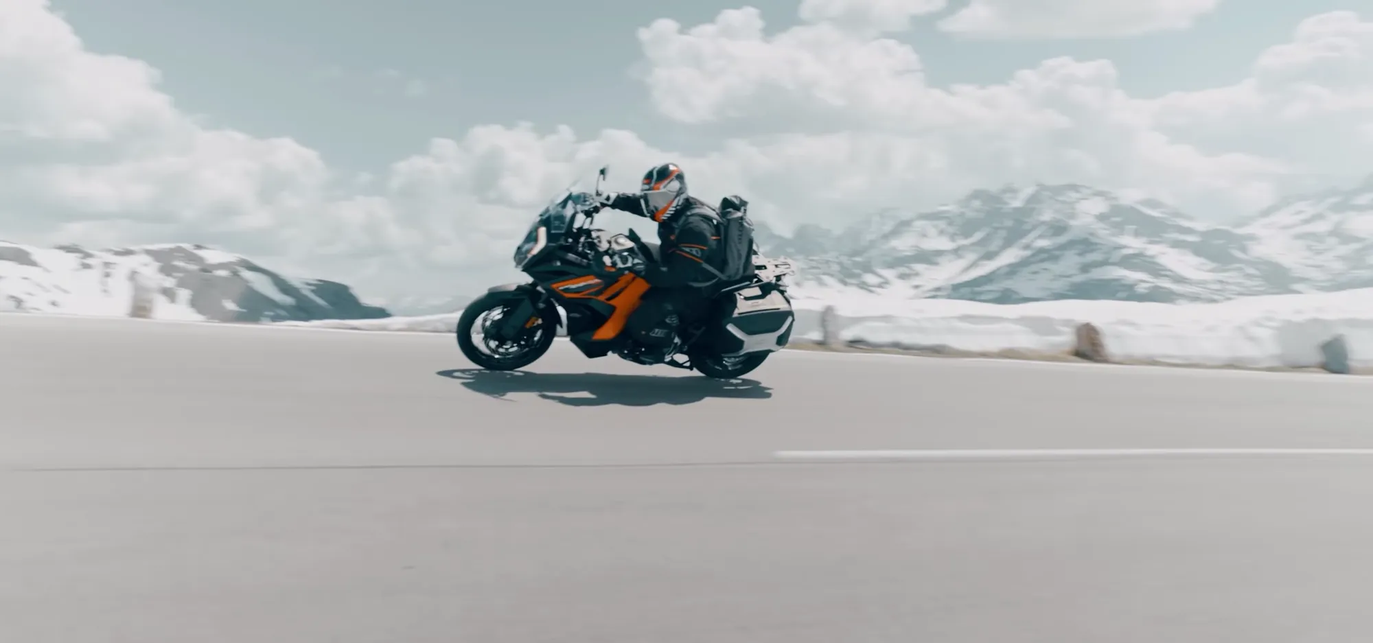 NastyNils riding a KTM 1290 Super Adventure R on a high-speed run across a flat plain with snow-capped mountains in the distance. Clear daylight, partly cloudy sky. The rider wears full black motorcycle gear and helmet, maintaining focus on the road. The bike displays KTM's characteristic orange and black livery. The landscape suggests alpine or high-altitude terrain with sparse vegetation.