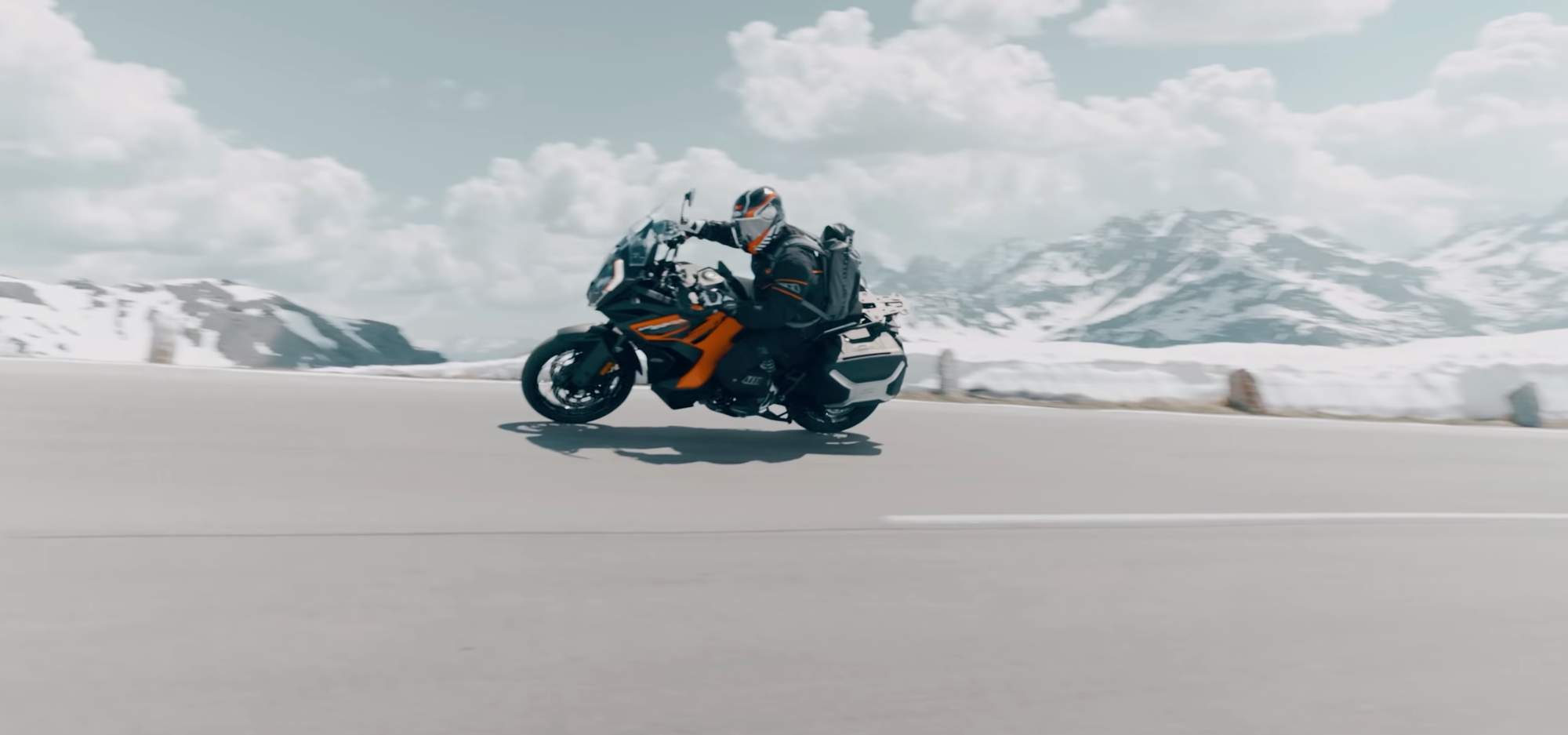 NastyNils riding a KTM 1290 Super Adventure R on a high-speed run across a flat plain with snow-capped mountains in the distance. Clear daylight, partly cloudy sky. The rider wears full black motorcycle gear and helmet, maintaining focus on the road. The bike displays KTM's characteristic orange and black livery. The landscape suggests alpine or high-altitude terrain with sparse vegetation.