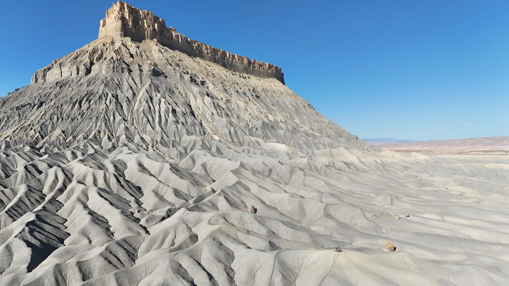Factory Butte in Utah's high desert badlands, captured in daylight under clear blue sky. The formation's distinctive multi-colored strata and steep erosional gullies dominate the frame. Arid terrain with minimal vegetation stretches across the foreground and background. Typical American Southwest landscape.