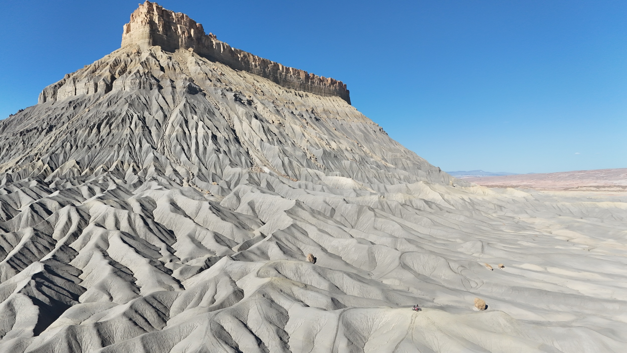 Factory Butte in Utah's high desert badlands, captured in daylight under clear blue sky. The formation's distinctive multi-colored strata and steep erosional gullies dominate the frame. Arid terrain with minimal vegetation stretches across the foreground and background. No motorcycle or person visible. Typical American Southwest landscape.