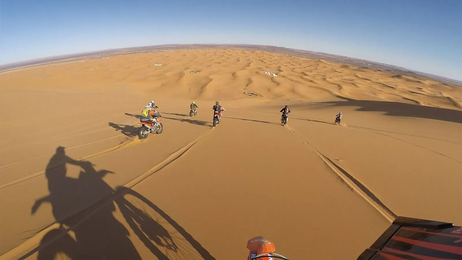 Handlebar view of an orange enduro in golden sand dunes, six riders ahead and the bike's shadow on the sand.