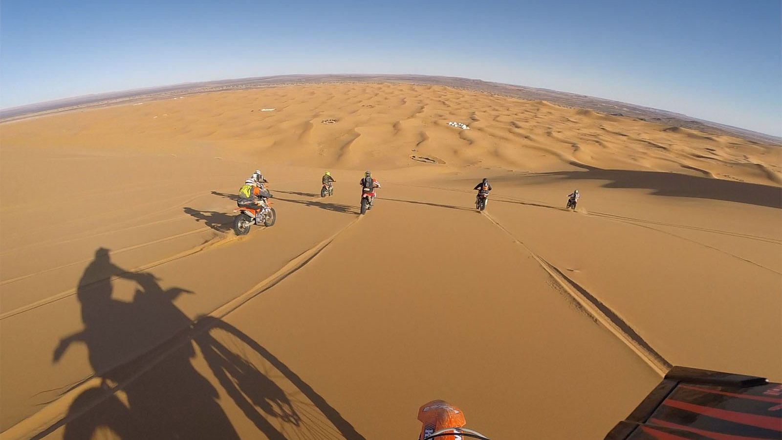 Handlebar view of an orange enduro in golden sand dunes, six riders ahead and the bike's shadow on the sand.