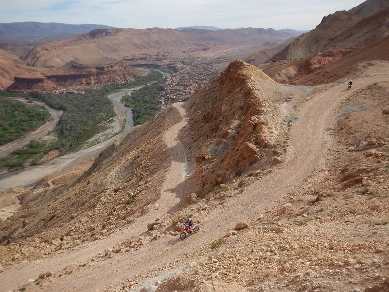 Two riders on a switchback gravel track on a barren mountainside above a green river valley with a village.