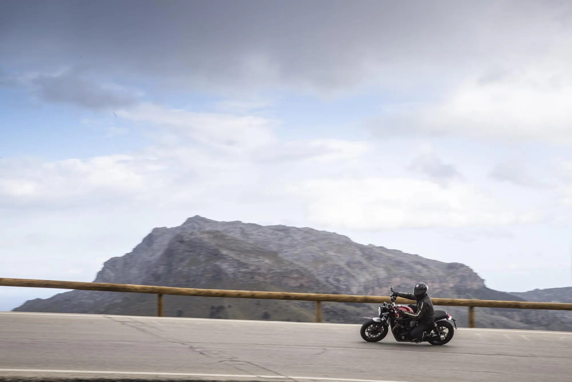 NastyNils riding a Triumph Speed Twin on an arid mountain pass, leaning into a curve beneath open sky. The red retro-classic motorcycle navigates winding asphalt with a yellow railing and desert mountain backdrop visible. Daylight, dry conditions, NastyNils in full black riding gear and helmet, focused on the road ahead. The scene captures the quintessential retro-modern riding experience of this parallel-twin classic.