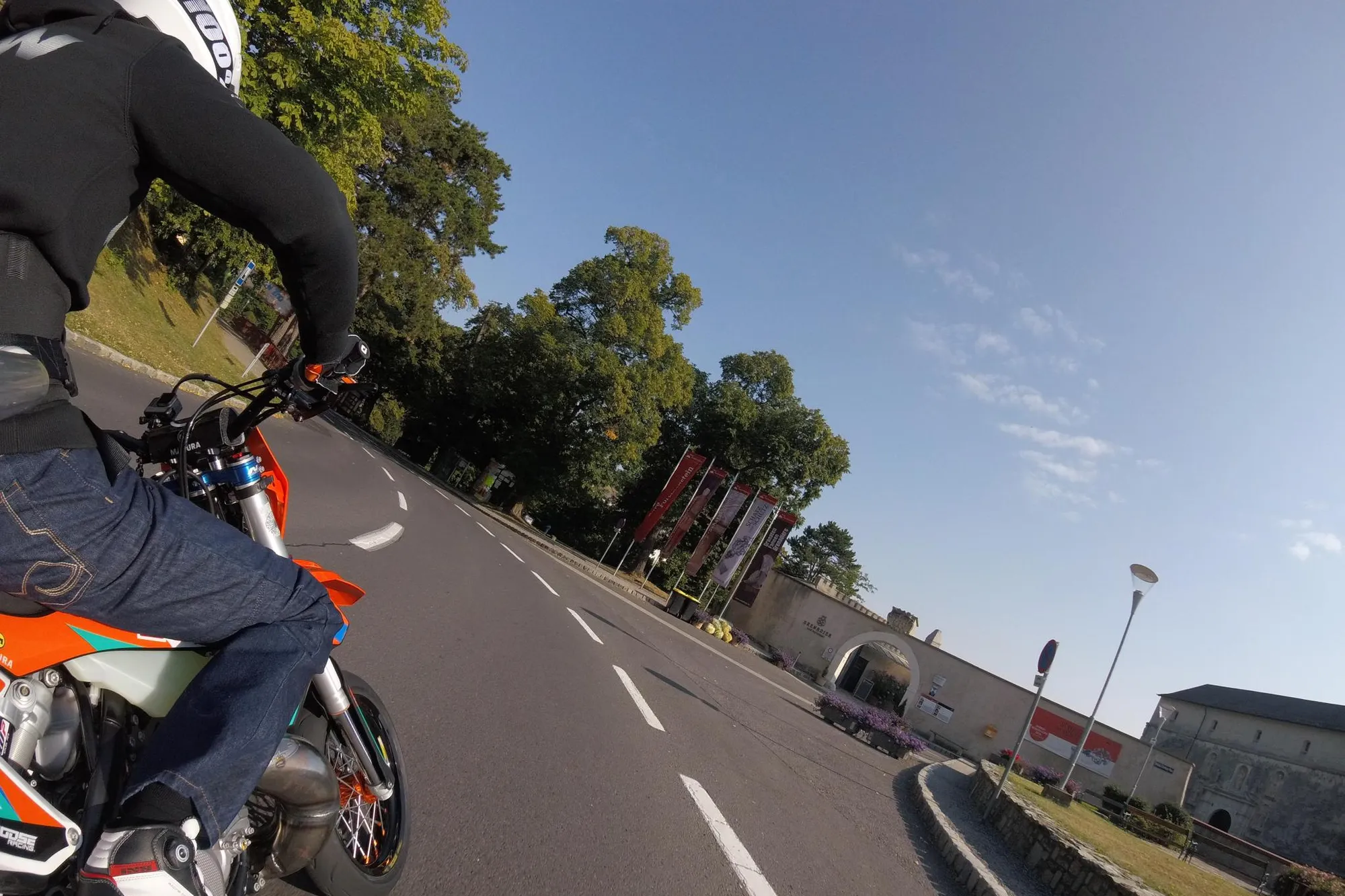 NastyNils riding a KTM 300 EXC in supermoto configuration on a public road near Forchtenstein, Austria, captured via onboard action camera. The rider leans through a right-hand corner wearing blue jeans, a dark jacket, and white helmet. The orange KTM with street-spec supermoto wheels and high-mounted exhaust is visible in the left foreground. A historic building consistent with Forchtenstein Castle and event banners appear to the right in background. Daylight, dry asphalt, clear sky.
