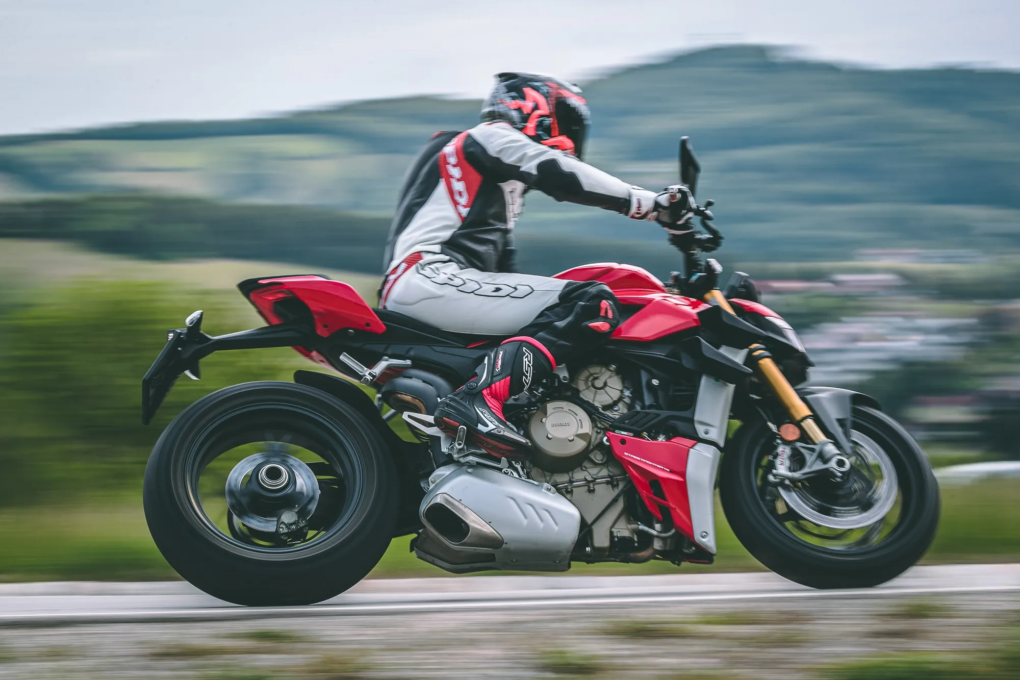 NastyNils riding a Ducati Streetfighter V4S on a winding rural road through rolling countryside. The rider is leaned aggressively through a corner, dressed in red and white leathers with full-face helmet. The V4S is shown in characteristic red and white livery with motion blur in the background indicating speed. Daylight, dry asphalt, dynamic action shot.