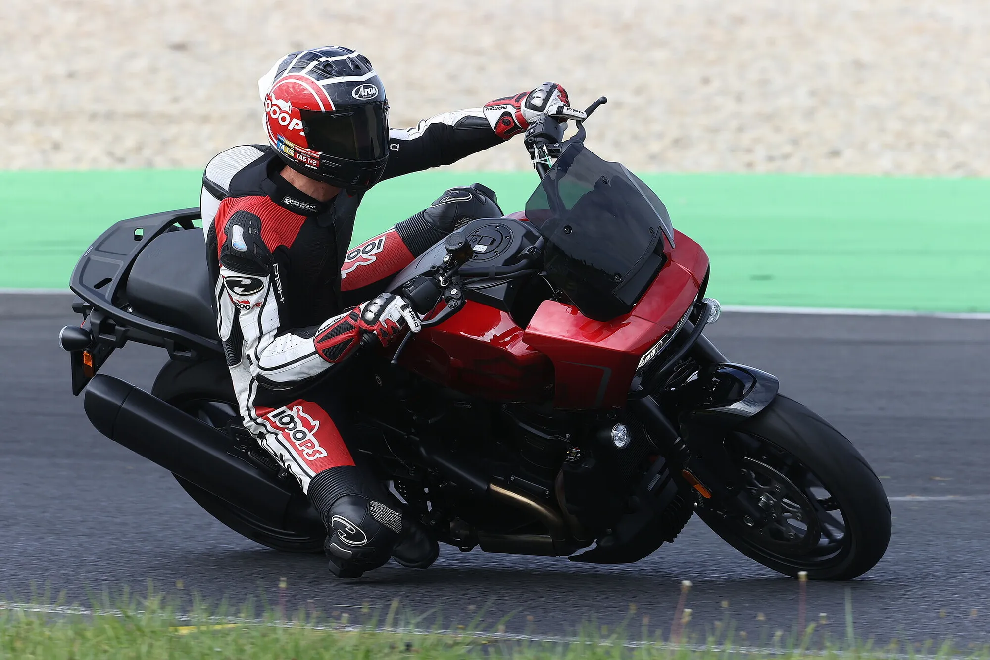 NastyNils on a Harley-Davidson Pan America 1250 ST with sidecar at a racetrack, waving to the camera. The motorcycle features red and black bodywork and is photographed in action on asphalt with a green safety barrier visible in the background. The rider wears a full-face Arai helmet and black leathers. Daylight, dry conditions, professional motorsports setting.