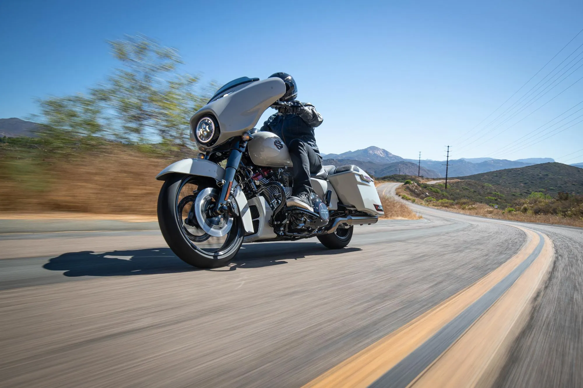 NastyNils riding a 2019 Harley-Davidson CVO Street Glide on a desert highway in California. The cruiser leans into a sweeping left turn under clear blue skies, with mountains and dry hills visible in the background. Chrome details, white wall tires, and the touring fairing catch the sunlight. Full leathers and helmet visible. Dry asphalt, daylight, smooth two-lane highway with yellow center line markings.