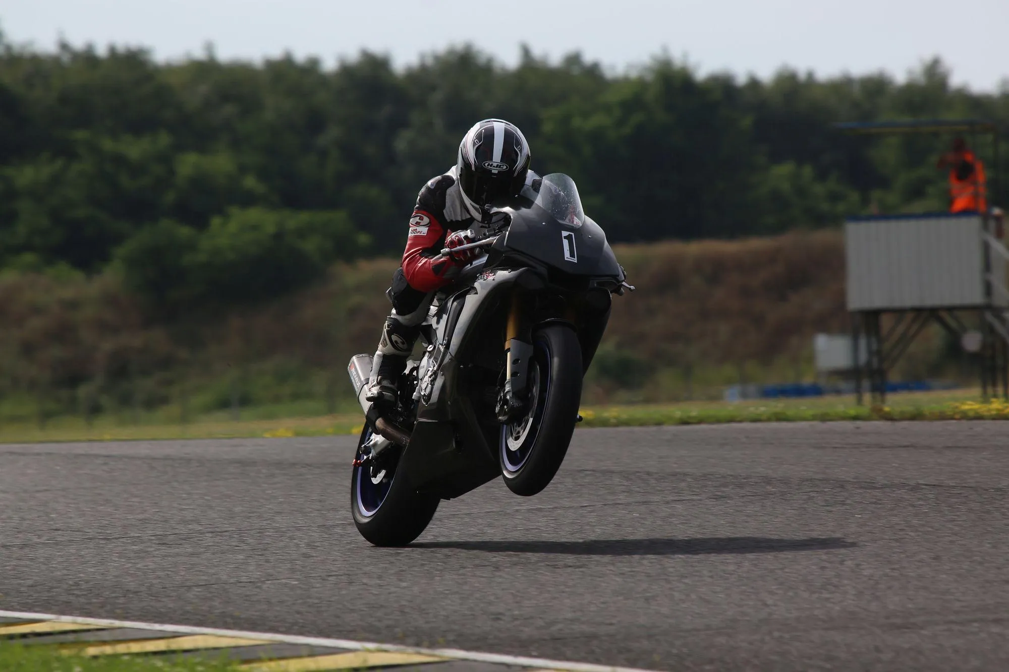 NastyNils performs a wheelie on a Yamaha YZF-R1M at the Pannoniaring racetrack in Hungary, 2019. The front wheel is fully airborne as the rider drives hard down the straight. Gold Öhlins inverted front forks and carbon-finish body panels identify the R1M variant. Rider wears full leathers in black, red, and white with an HJC full-face helmet. A trackside marshal post with safety crew in orange high-vis is visible in the background.