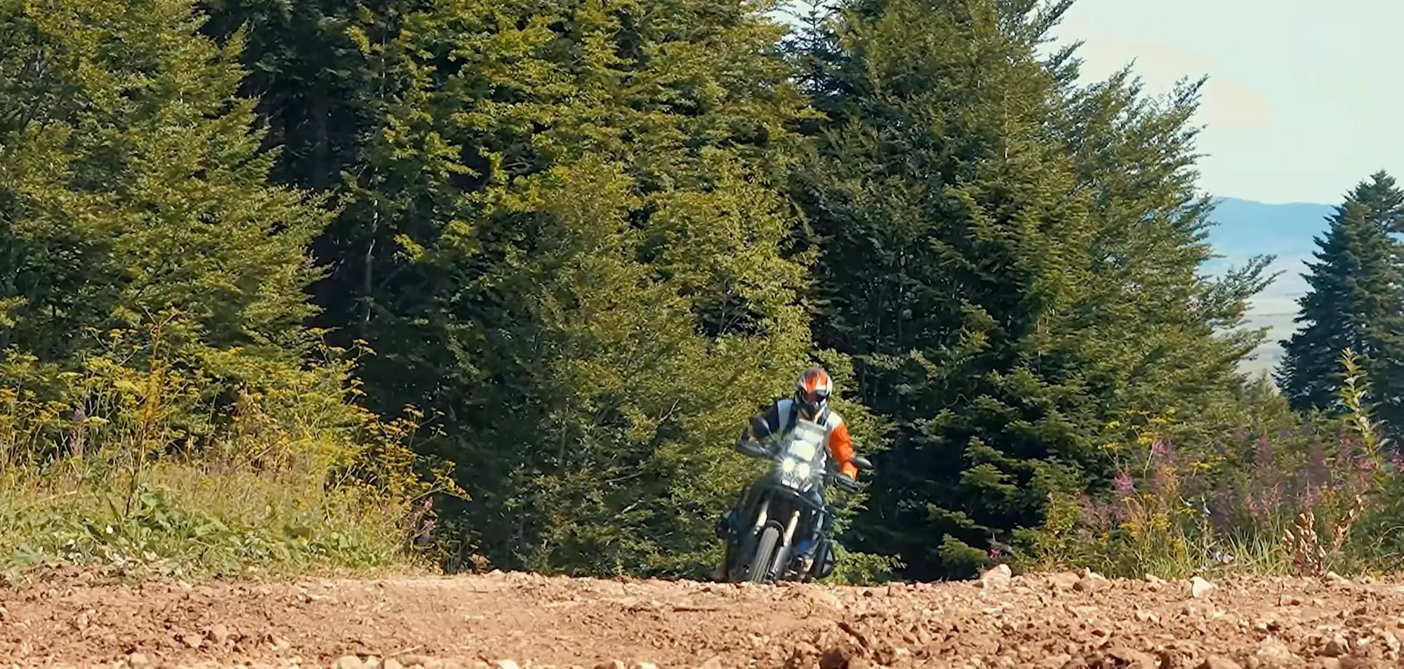 NastyNils riding a Yamaha Tenere 700 on a gravel rally route in Bosnia, leaning into a gentle turn through forested terrain. The cream-colored adventure bike is in motion on the dirt surface with dense pine forest visible in the background. Daylight conditions, NastyNils in full protective gear.