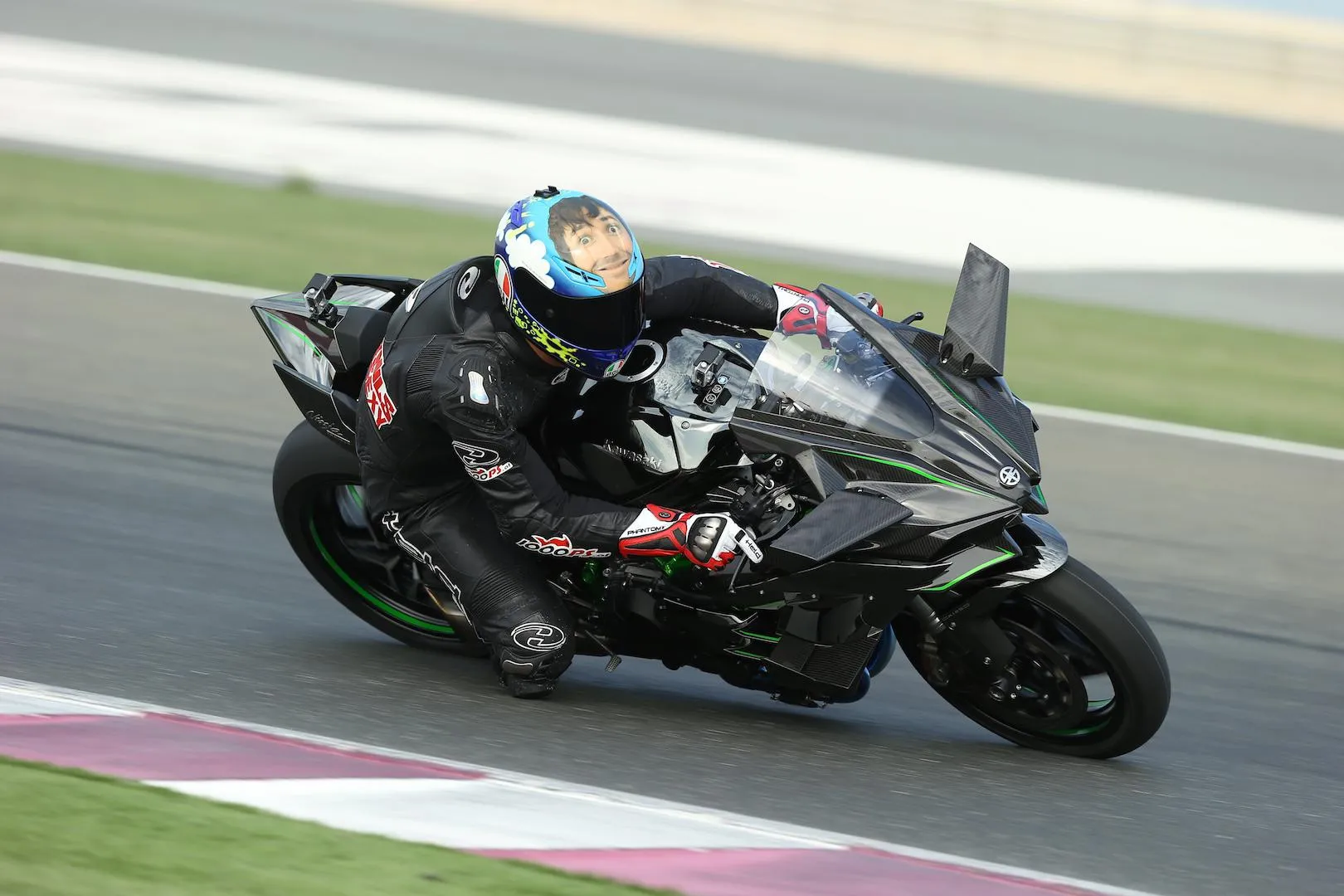 NastyNils riding a Kawasaki Ninja H2 family motorcycle at speed through a left-hand corner on an asphalt racing circuit. The bike's large swept aerodynamic winglets and carbon-fiber fairings are clearly visible. Rider wears black 1000PS-branded leather suit and a blue helmet with a custom illustrated cartoon-face graphic. Track features pink and green kerbing consistent with a professional international circuit, likely Middle East.