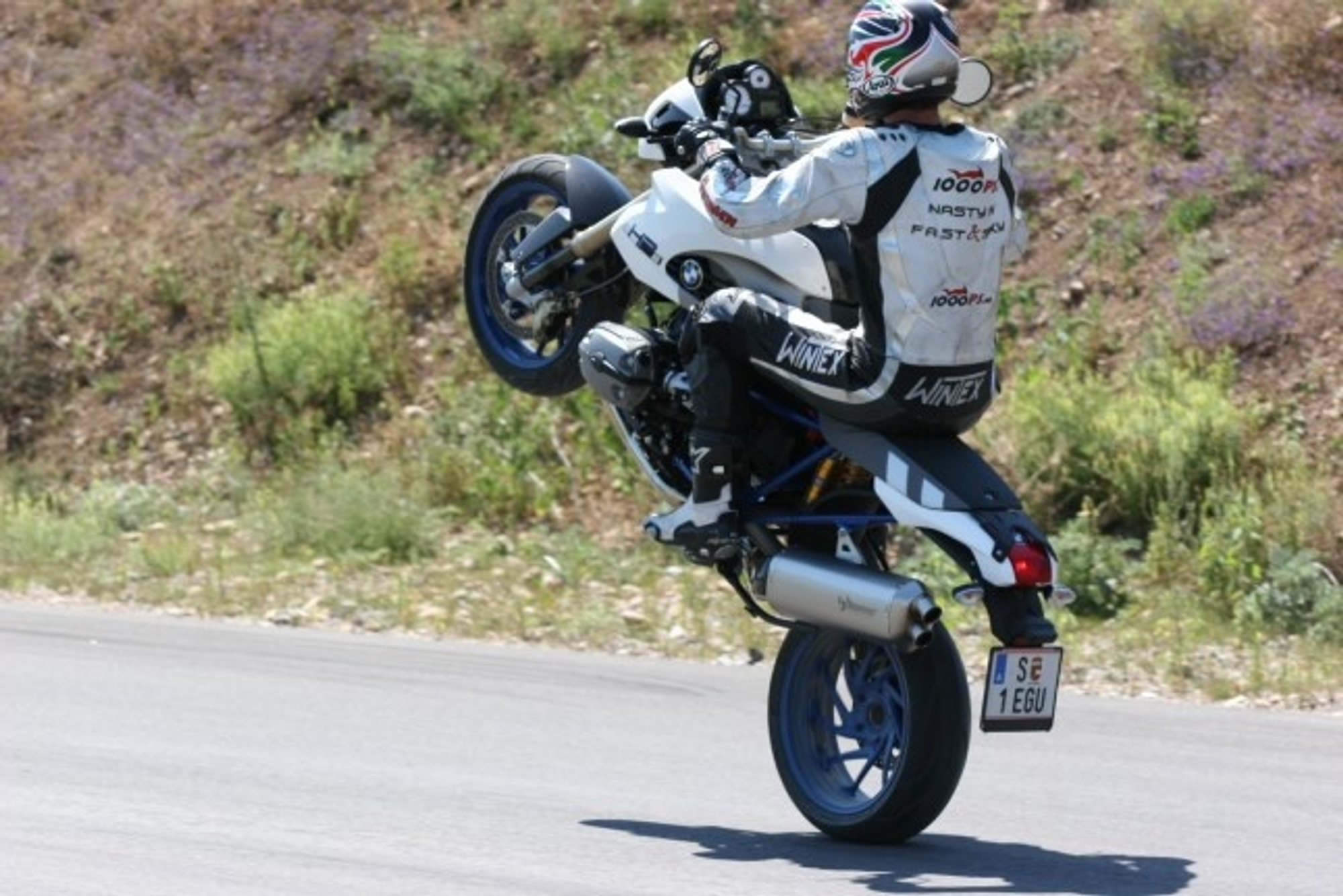 NastyNils performs a controlled wheelie on a white-and-black BMW HP2 Megamoto supermoto on a rural asphalt road, with scrubby hills and sparse vegetation visible in the background. The bike balances on its front wheel in an extended stunt. Clear daylight, dry conditions. NastyNils wears black-and-white leather riding gear and a white graphic race helmet. Swedish license plate visible.