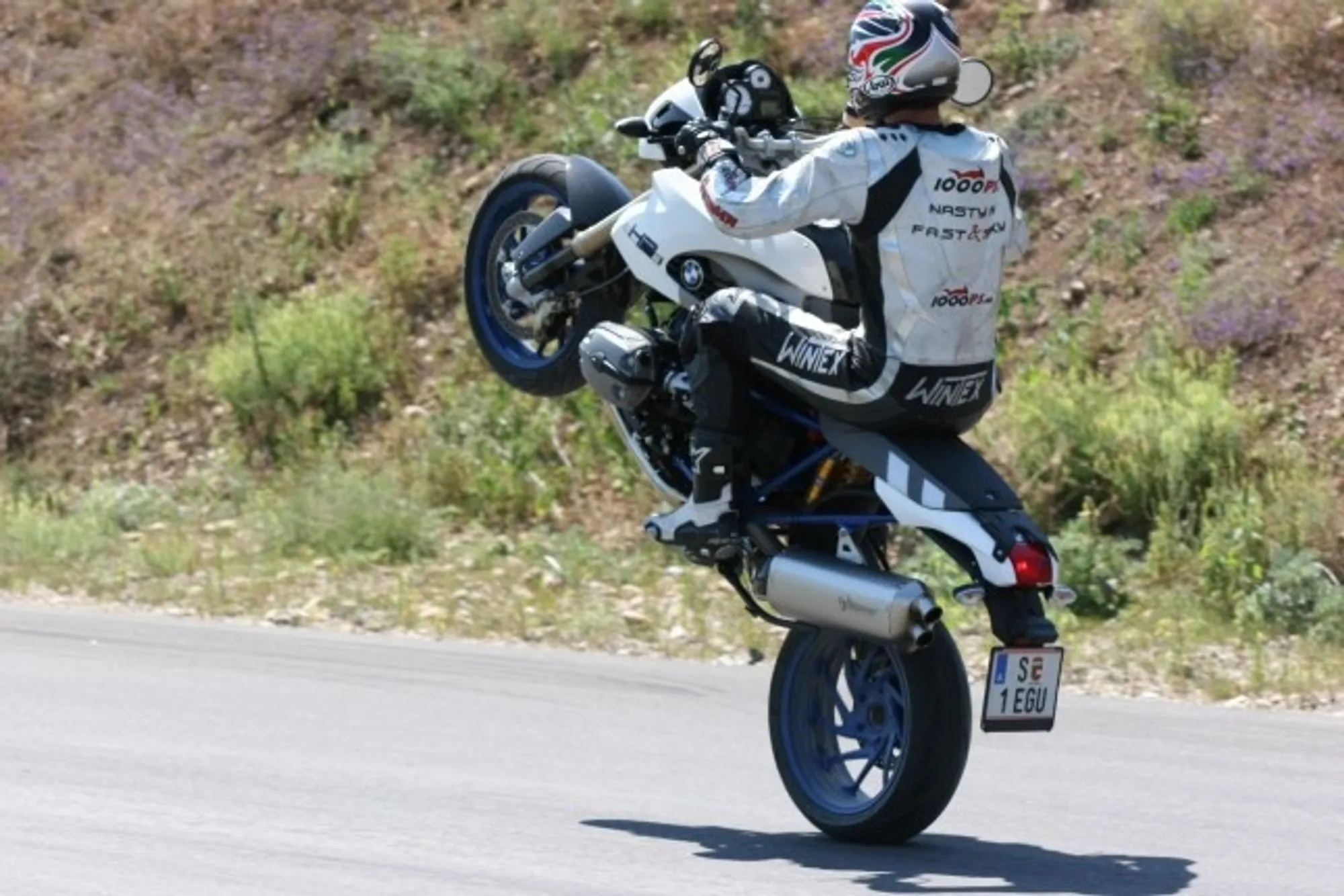 NastyNils performs a controlled wheelie on a white-and-black BMW HP2 Megamoto supermoto on a rural asphalt road, with scrubby hills and sparse vegetation visible in the background. The bike balances on its front wheel in an extended stunt. Clear daylight, dry conditions. NastyNils wears black-and-white leather riding gear and a white graphic race helmet. Swedish license plate visible.