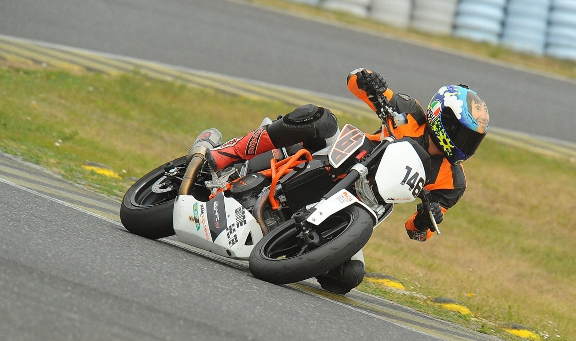 NastyNils riding a KTM 690 Duke on a racetrack in aggressive cornering attack, leaning deep with near-kneedown body positioning. The race-configured 690 Duke displays white-and-orange livery with race number 146. Daylight, dry asphalt circuit with grass run-off area and tire barriers visible. Rider in red/black racing leathers and multicolor race helmet, fully committed to the corner.