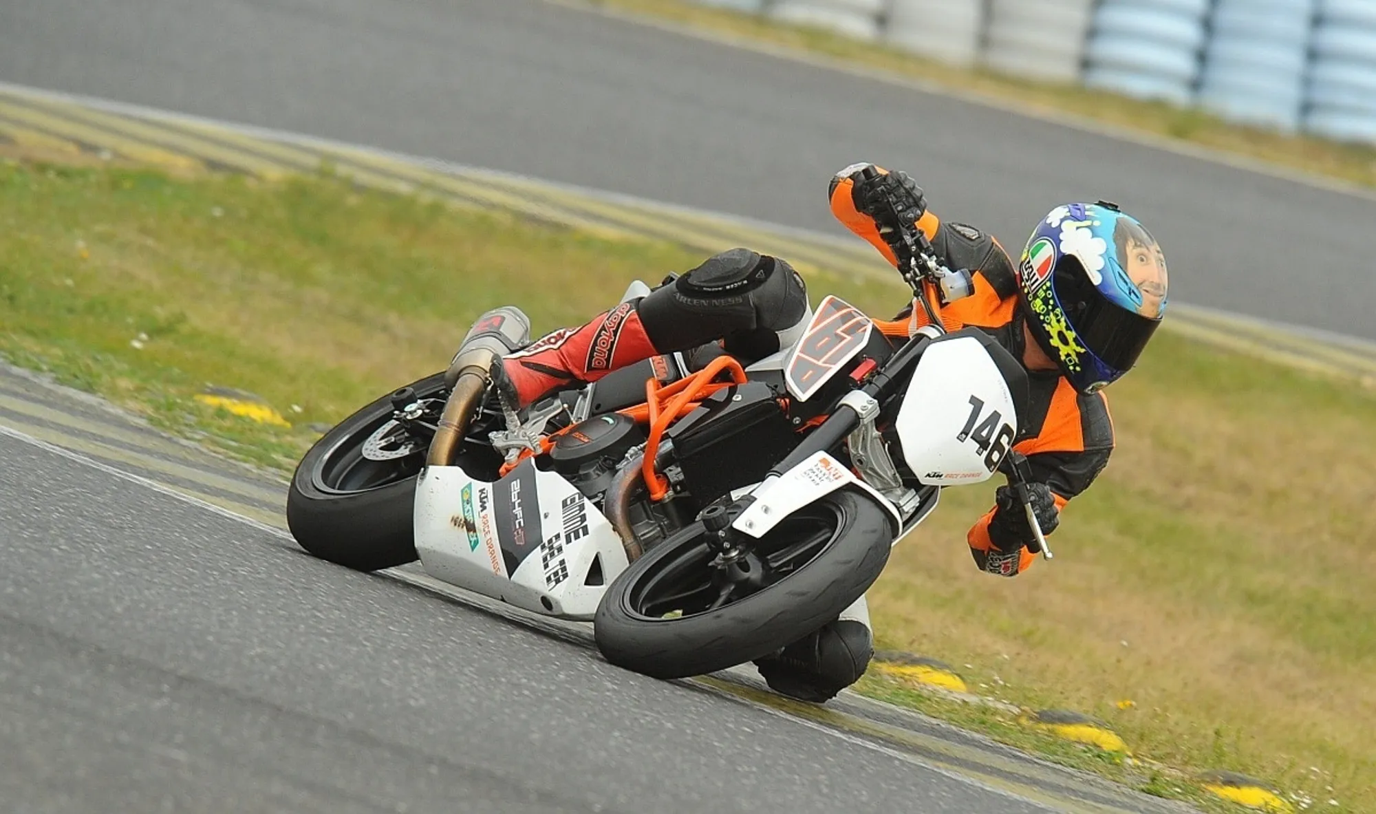 NastyNils riding a KTM 690 Duke on a racetrack in aggressive cornering attack, leaning deep with near-kneedown body positioning. The race-configured 690 Duke displays white-and-orange livery with race number 146. Daylight, dry asphalt circuit with grass run-off area and tire barriers visible. Rider in red/black racing leathers and multicolor race helmet, fully committed to the corner.