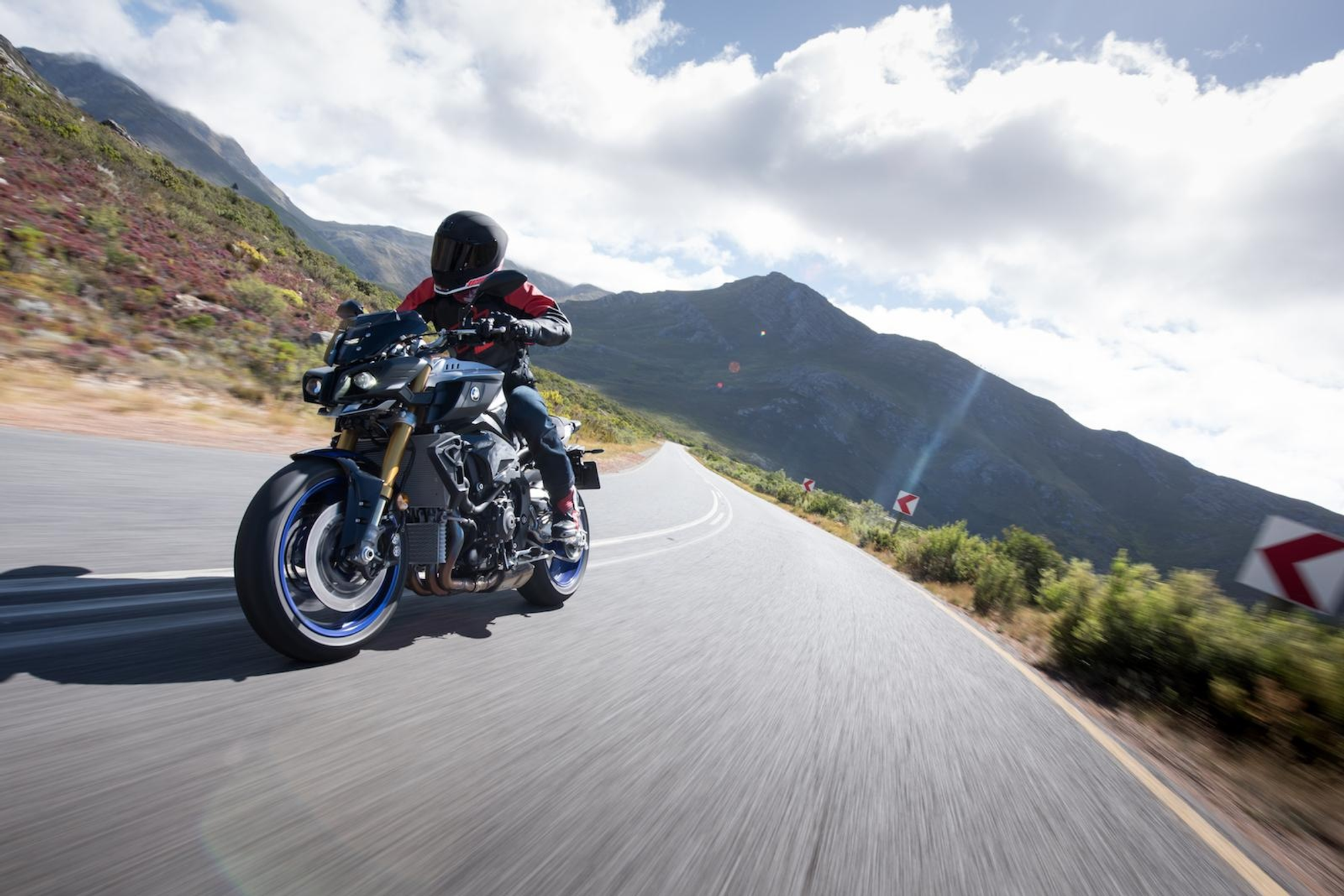 NastyNils riding a Yamaha MT-10 SP on a winding mountain road, leaning through a right-hand bend at speed. Motion blur on the road surface confirms high-speed tracking shot. Mountain ridgeline and partly cloudy sky dominate the background. Rider wears a black full-face helmet and red/black riding jacket. Blue forged aluminum wheels and Öhlins front fork confirm SP specification. Dry asphalt, daylight, open landscape consistent with South Africa press-launch setting.