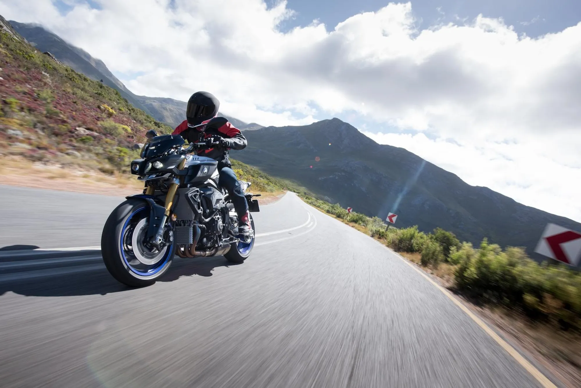NastyNils riding a Yamaha MT-10 SP on a winding mountain road, leaning through a right-hand bend at speed. Motion blur on the road surface confirms high-speed tracking shot. Mountain ridgeline and partly cloudy sky dominate the background. Rider wears a black full-face helmet and red/black riding jacket. Blue forged aluminum wheels and Öhlins front fork confirm SP specification. Dry asphalt, daylight, open landscape consistent with South Africa press-launch setting.