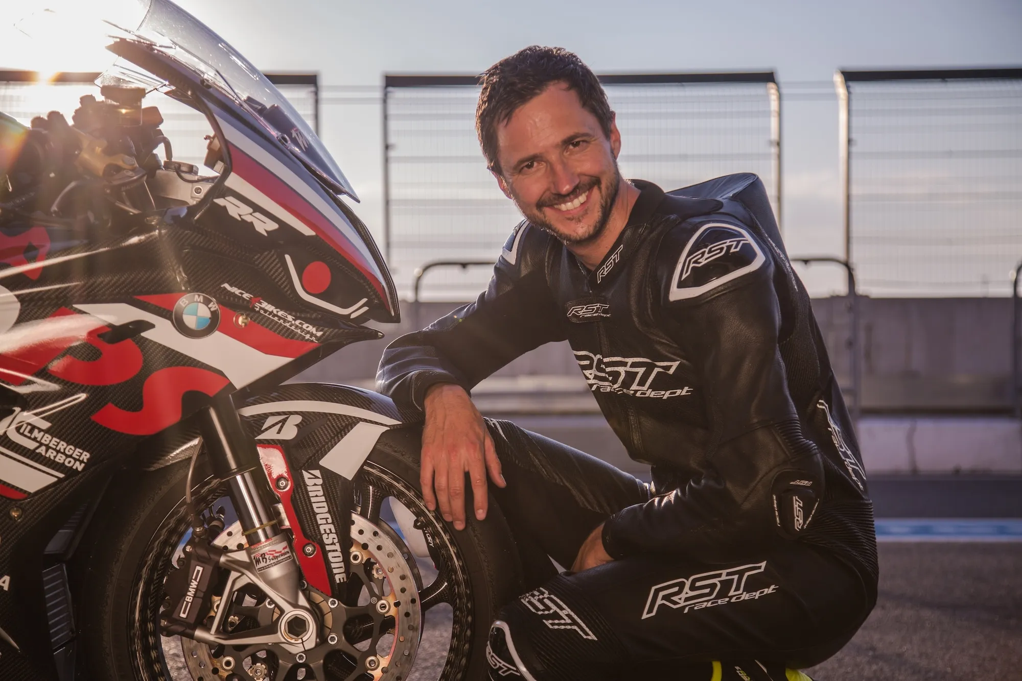 NastyNils posing next to a BMW S1000RR sportsbike in a track paddock setting, wearing full black racing leathers with RST branding. The bike displays classic red, white, and black livery. Golden hour lighting illuminates the scene. High-quality press photography with blurred safety track fencing in the background. Relaxed, confident posture with hand resting on the bike.