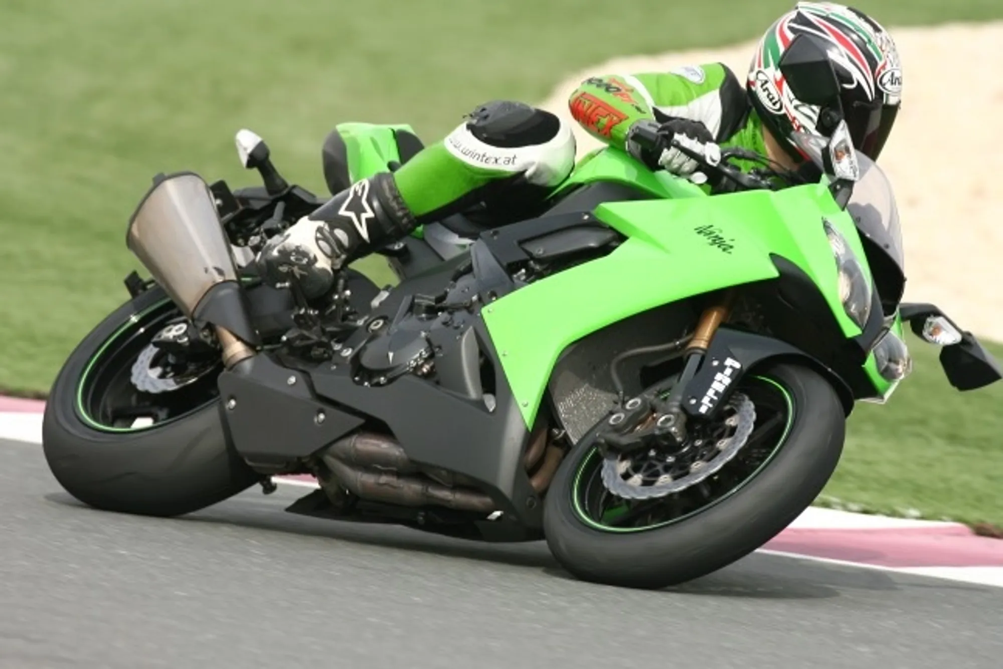 NastyNils riding a Kawasaki ZX10R in striking lime-green livery on a professional racetrack, leaning through an aggressive high-speed right-hander with knee-down lean angle. Full leathers, full-face helmet. Tarmac track with painted curbing and green grass runoff visible. Daylight, clear conditions, professional track environment.