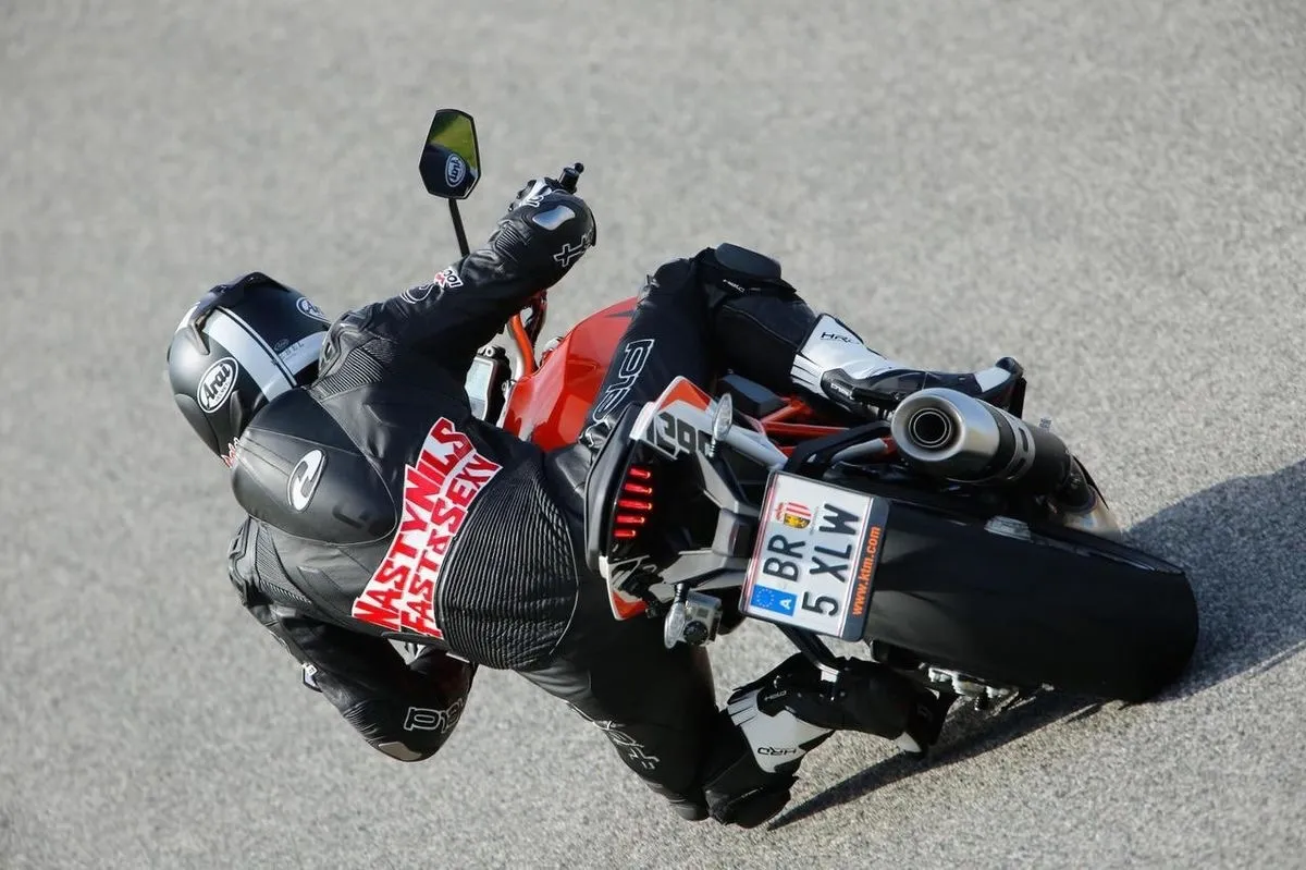 NastyNils riding a KTM Super Duke R in a high-lean cornering action at a paved circuit. The rider wears full black leather suit with protective armor and full-face helmet, knee down on dry asphalt. Daylight conditions. The aggressive lean angle and body positioning demonstrate the bike's track-oriented handling characteristics.