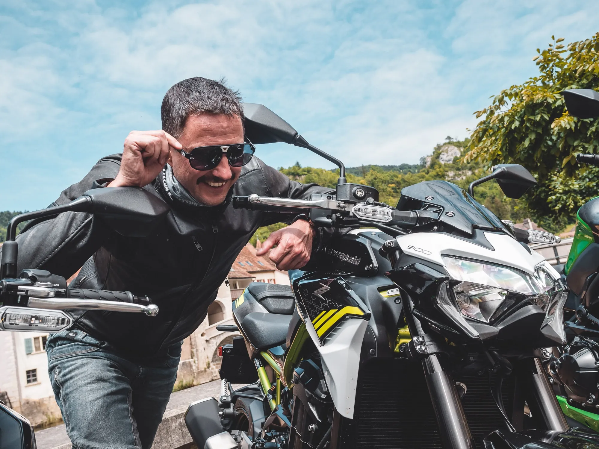 NastyNils poses with a Kawasaki Z900 naked motorcycle in an outdoor staging area. He wears dark sunglasses, black leather riding jacket, and jeans, smiling at the camera with a relaxed expression. The Z900 is displayed in static pose, facing the camera. Clear blue sky with green trees visible in background. Daylight photography, casual portrait style typical of personal bike shots.