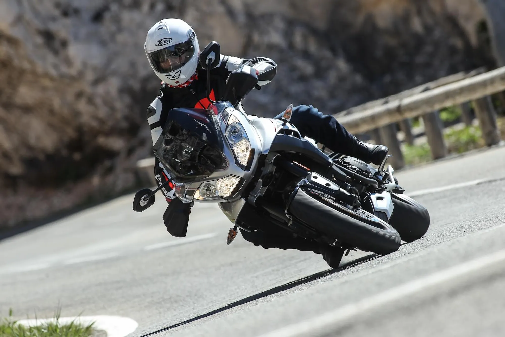 NastyNils corners aggressively on a Triumph Tiger Sport on a winding mountain road, leaning deeply into a left-hander with the footrests scraping the asphalt. Rocky cliff face and metal guardrail visible in the background. Daylight, dry road surface. NastyNils wears a white X-lite full-face helmet, black-and-red textile jacket, and dark riding trousers.