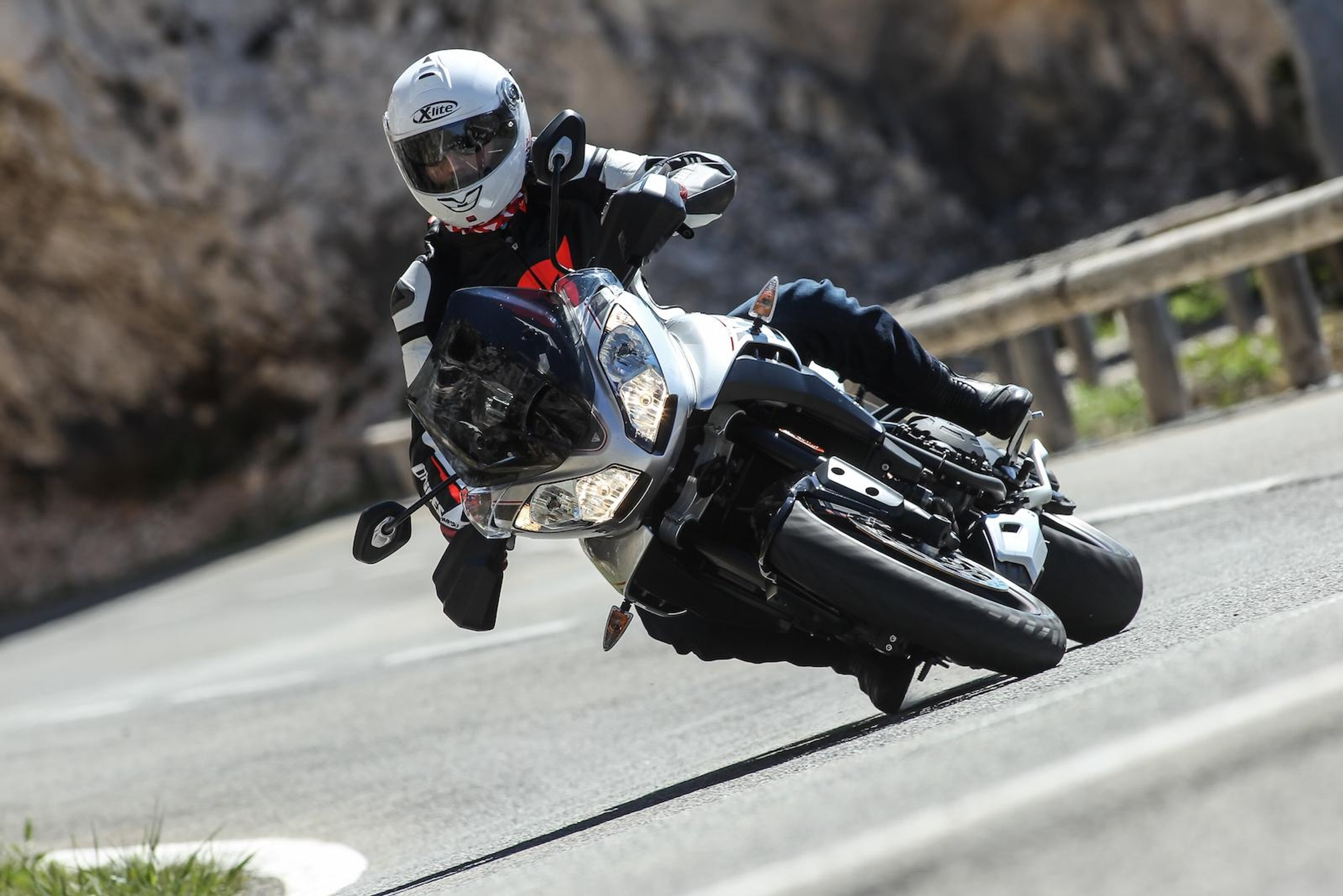NastyNils corners aggressively on a Triumph Tiger Sport on a winding mountain road, leaning deeply into a left-hander with the footrests scraping the asphalt. Rocky cliff face and metal guardrail visible in the background. Daylight, dry road surface. NastyNils wears a white X-lite full-face helmet, black-and-red textile jacket, and dark riding trousers.