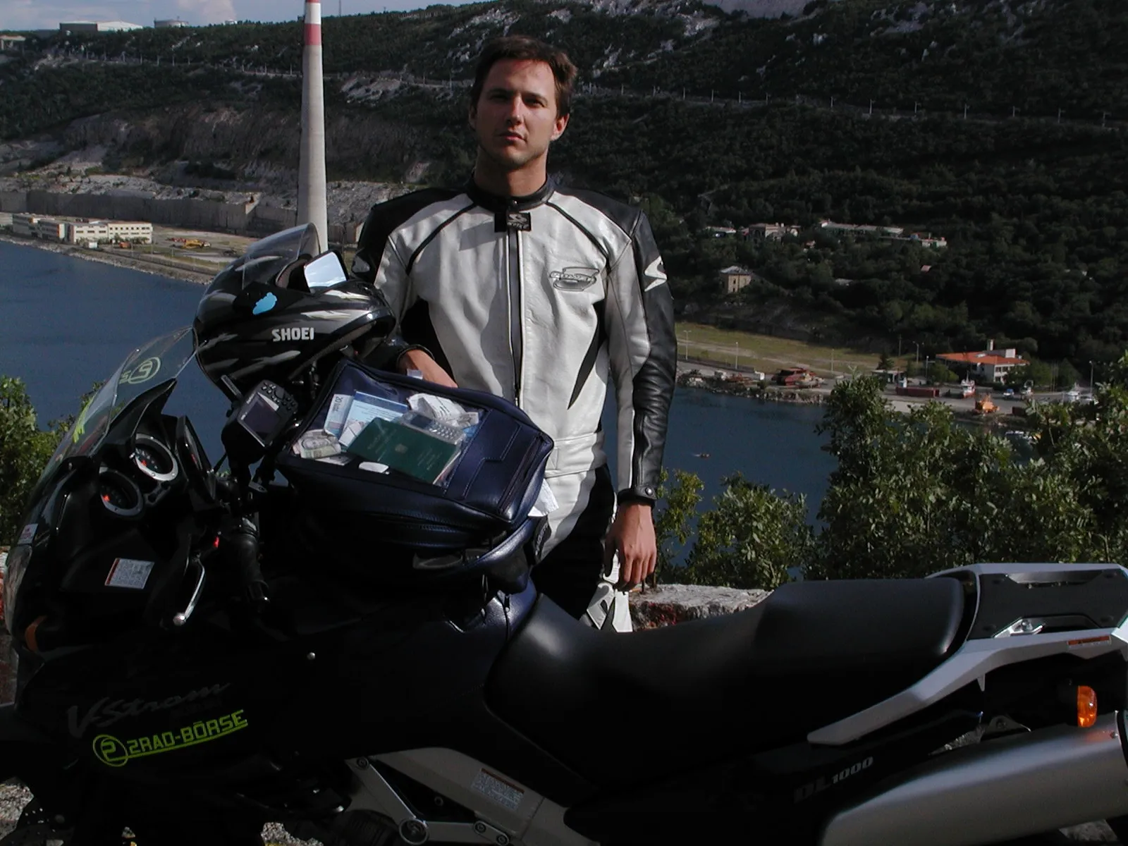 NastyNils poses with a Suzuki V-Strom adventure tourer on a coastal cliff overlooking a Croatian bay. The early-2000s adventure bike is equipped with touring luggage. NastyNils wears a mesh sports jacket and Shoei helmet. Limestone cliffs, forested hills, and Mediterranean coastline visible in background. Clear daylight, calm water. Scene captures adventure-touring preparation from a 2002 Croatia trip.