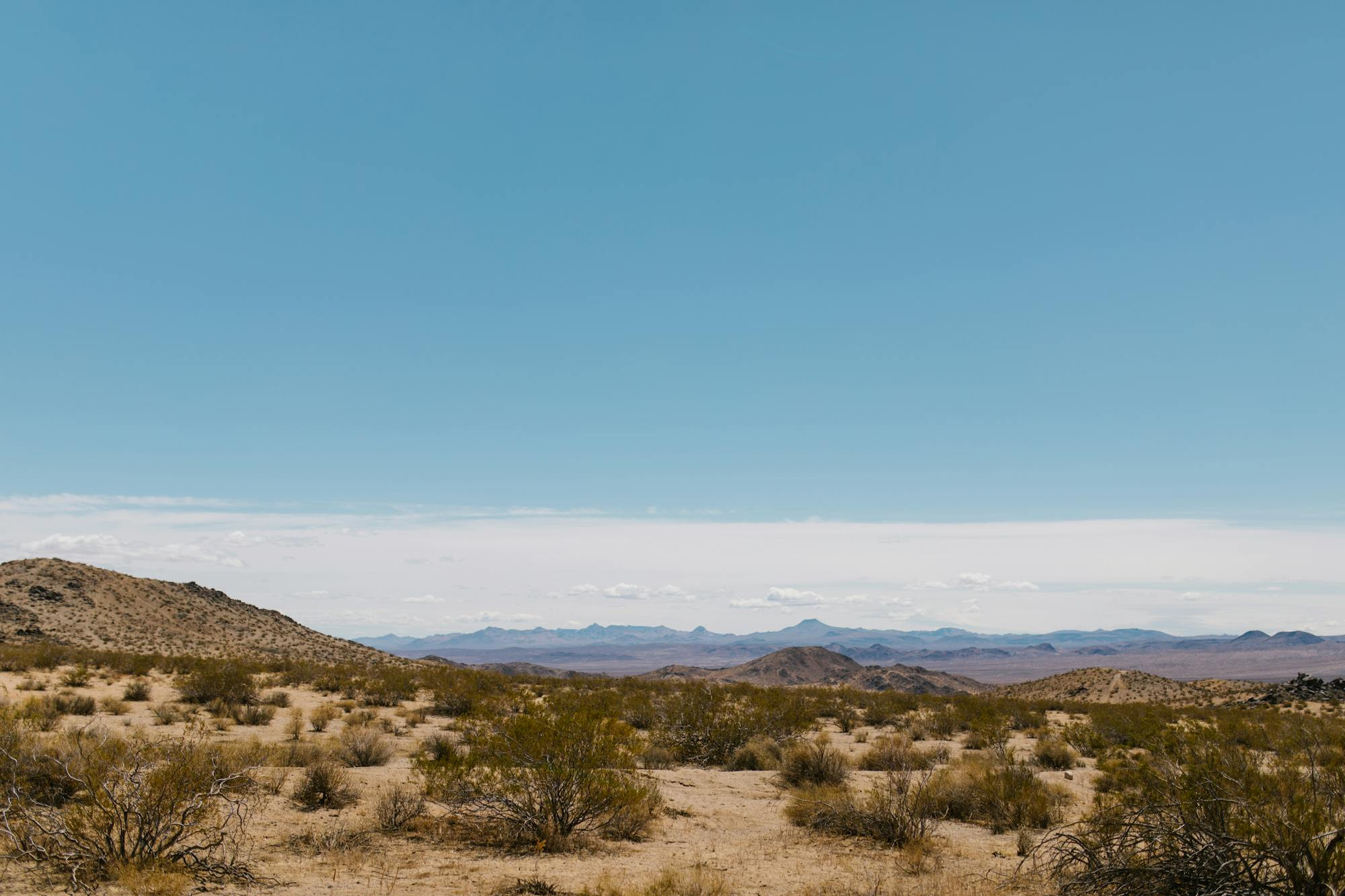 Wide desert landscape at Ocotillo Wells, California, showing sandy arid terrain with sparse desert scrub in the foreground, a rocky hill to the left, and a distant mountain range along the horizon. Clear blue sky with a thin band of light clouds at mid-height. No motorcycle or person visible. Pexels stock photograph by RDNE Stock Project, likely used as a mood or location establishing shot for an electric trail-riding editorial.