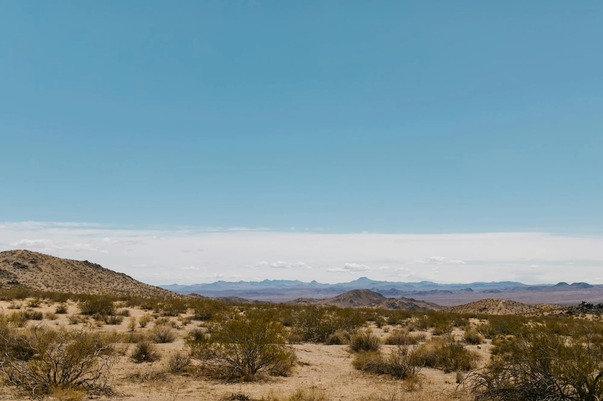 Wide desert landscape at Ocotillo Wells, California, showing sandy arid terrain with sparse desert scrub in the foreground, a rocky hill to the left, and a distant mountain range along the horizon. Clear blue sky with a thin band of light clouds at mid-height. No motorcycle or person visible. Pexels stock photograph by RDNE Stock Project, likely used as a mood or location establishing shot for an electric trail-riding editorial.