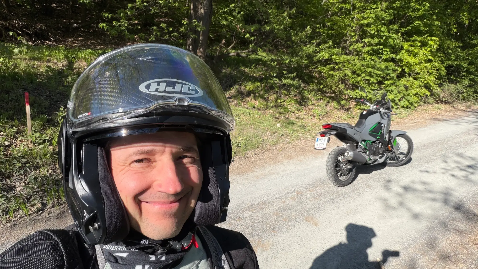 Nils Mueller taking a selfie with the 2026 Kawasaki KLE 500 on a gravel road in Lower Austria, full riding gear, lush forest in the background.
