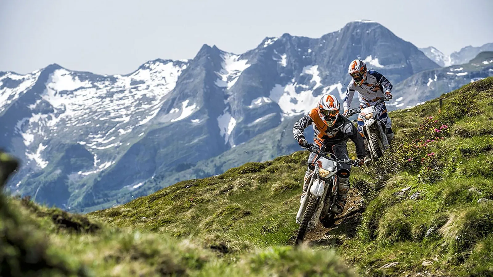 Two enduro riders on a grassy alpine slope beneath rocky peaks with patches of snow.