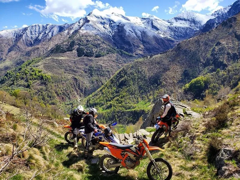 Enduro riders and parked dirt bikes above a green alpine valley with snow on the peaks.