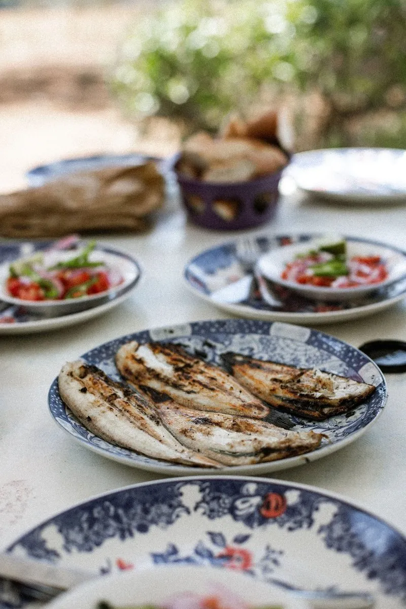 Grilled sardines on a plate with salads and bread on an outdoor table.