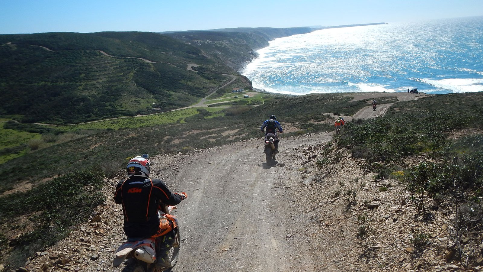 Four riders on enduros descending a gravel track on a scrubby slope toward the Atlantic coast, with a long beach and surf below.