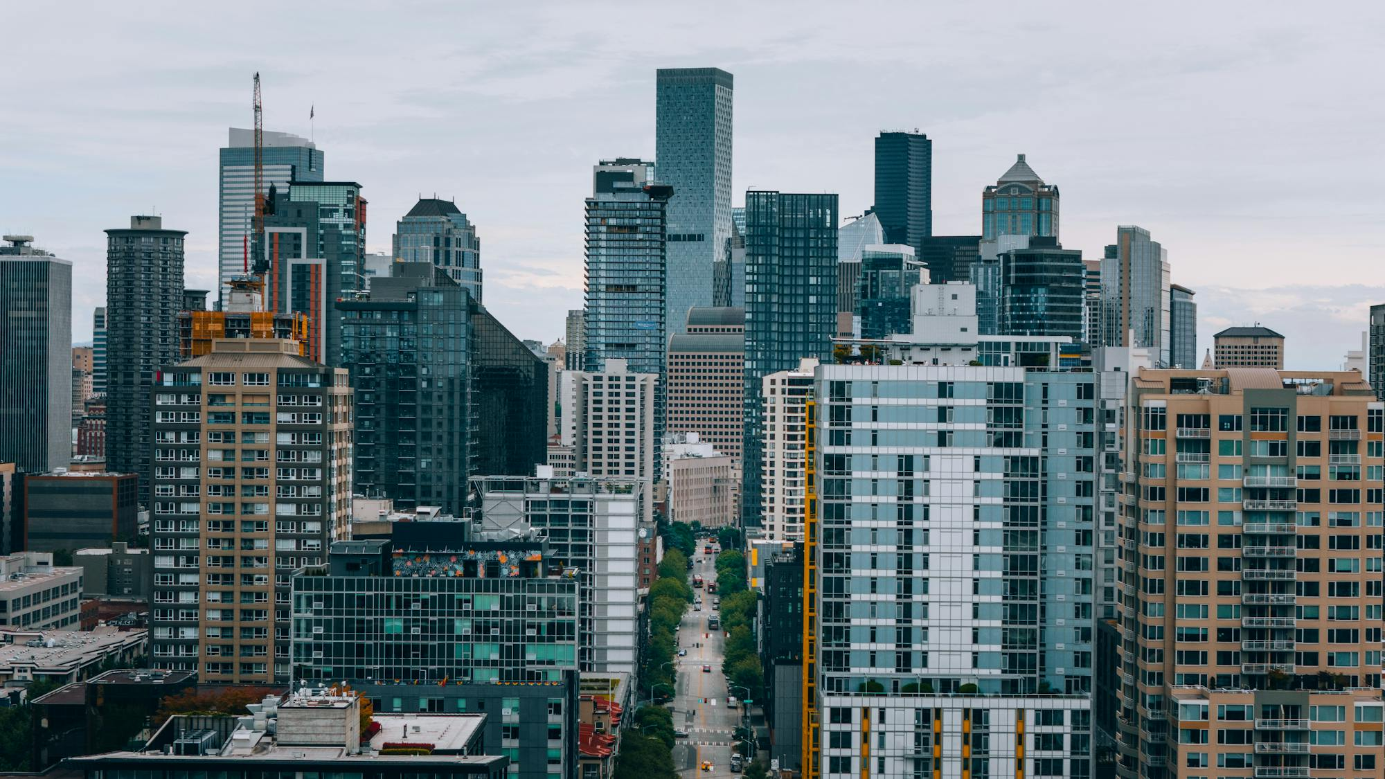Elevated aerial view of Seattle's downtown skyline looking toward the city core along a dense urban street canyon. A mix of glass-and-steel skyscrapers and mid-rise residential towers fills the frame. A construction crane is active on the left. The street below carries light vehicle traffic flanked by mature street trees. Overcast daylight, no motorcycle or person visible. Pexels stock photograph credited to Josh Hild.