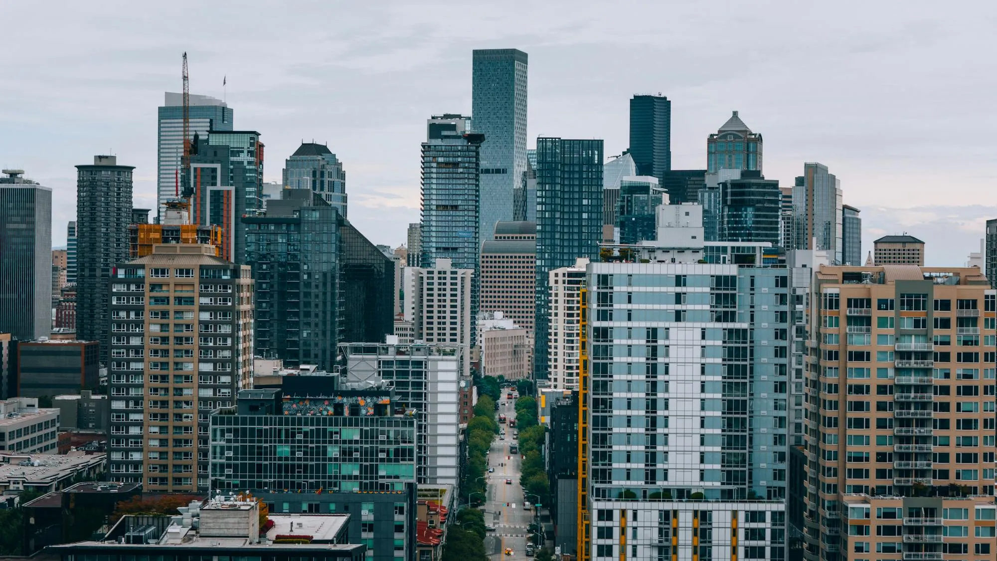Elevated aerial view of Seattle's downtown skyline looking toward the city core along a dense urban street canyon. A mix of glass-and-steel skyscrapers and mid-rise residential towers fills the frame. A construction crane is active on the left. The street below carries light vehicle traffic flanked by mature street trees.