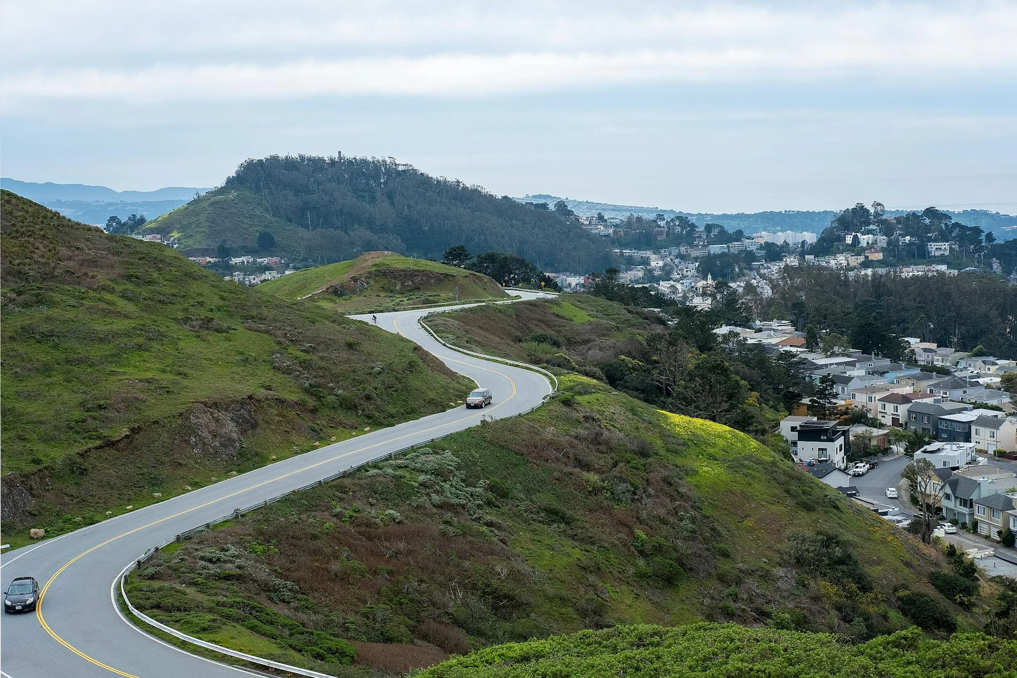 Aerial view of a winding asphalt road traversing rolling green hills in the Bay Area, likely Skyline Boulevard. The road curves through lush grassland with residential development visible in the distance. Clear weather, daylight, no motorcycle or rider visible.