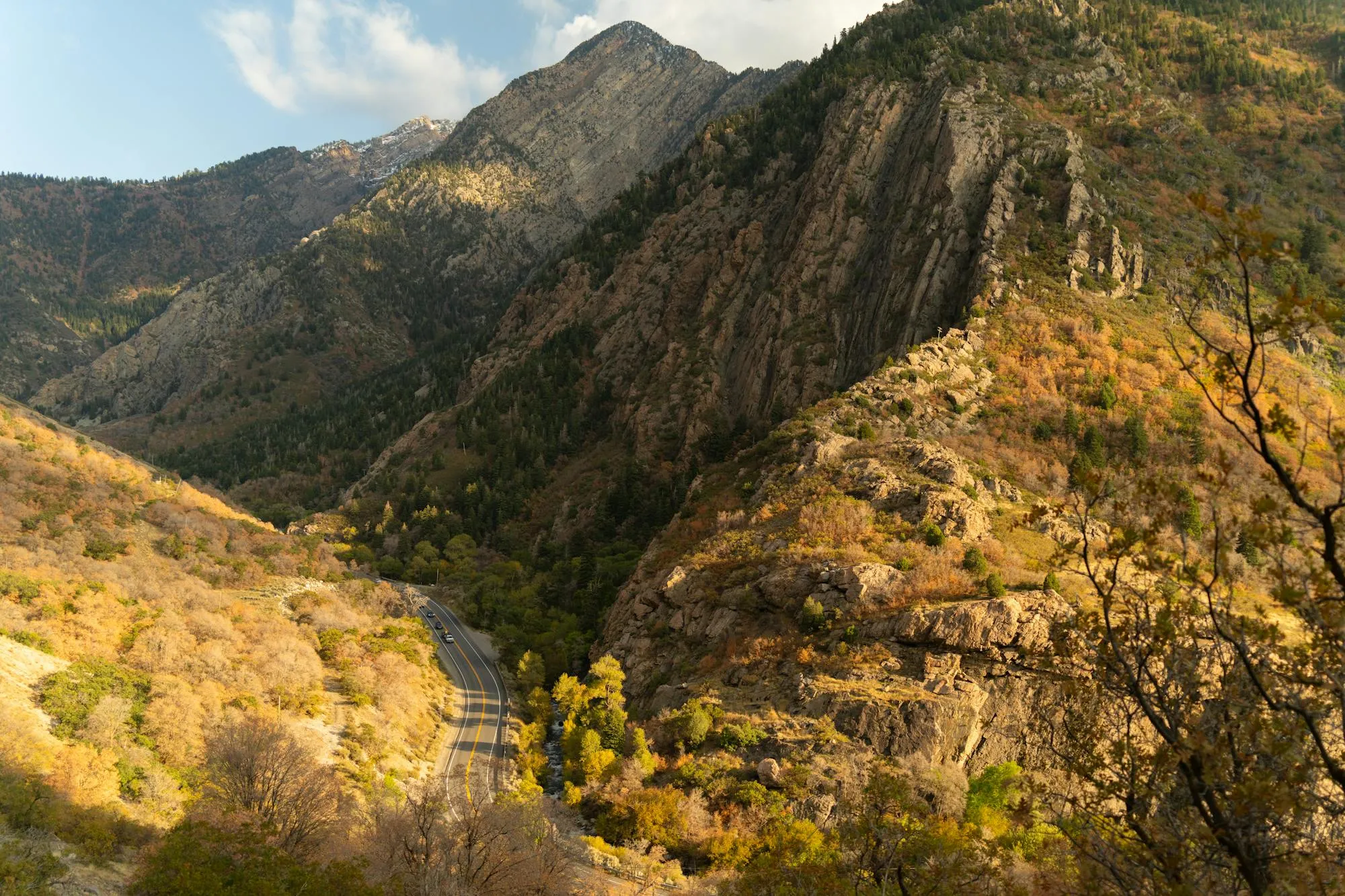 An elevated view of a deep autumn canyon, likely Big Cottonwood Canyon, Utah. Steep rocky cliff faces and forested mountain ridges frame a narrow valley where a winding two-lane road passes below. Deciduous trees display full autumn color — gold, orange, and amber — interspersed with green conifers on the steep slopes. A single dark vehicle is visible far below on the road. Snow-dusted mountain peaks rise in the background under a partly cloudy sky.