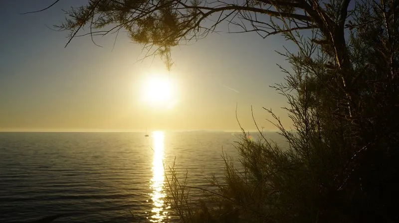 Sunset over the Adriatic Sea near Primosten, Croatia, with golden hour light bathing calm water in warm tones, a small sailboat on the distant horizon.