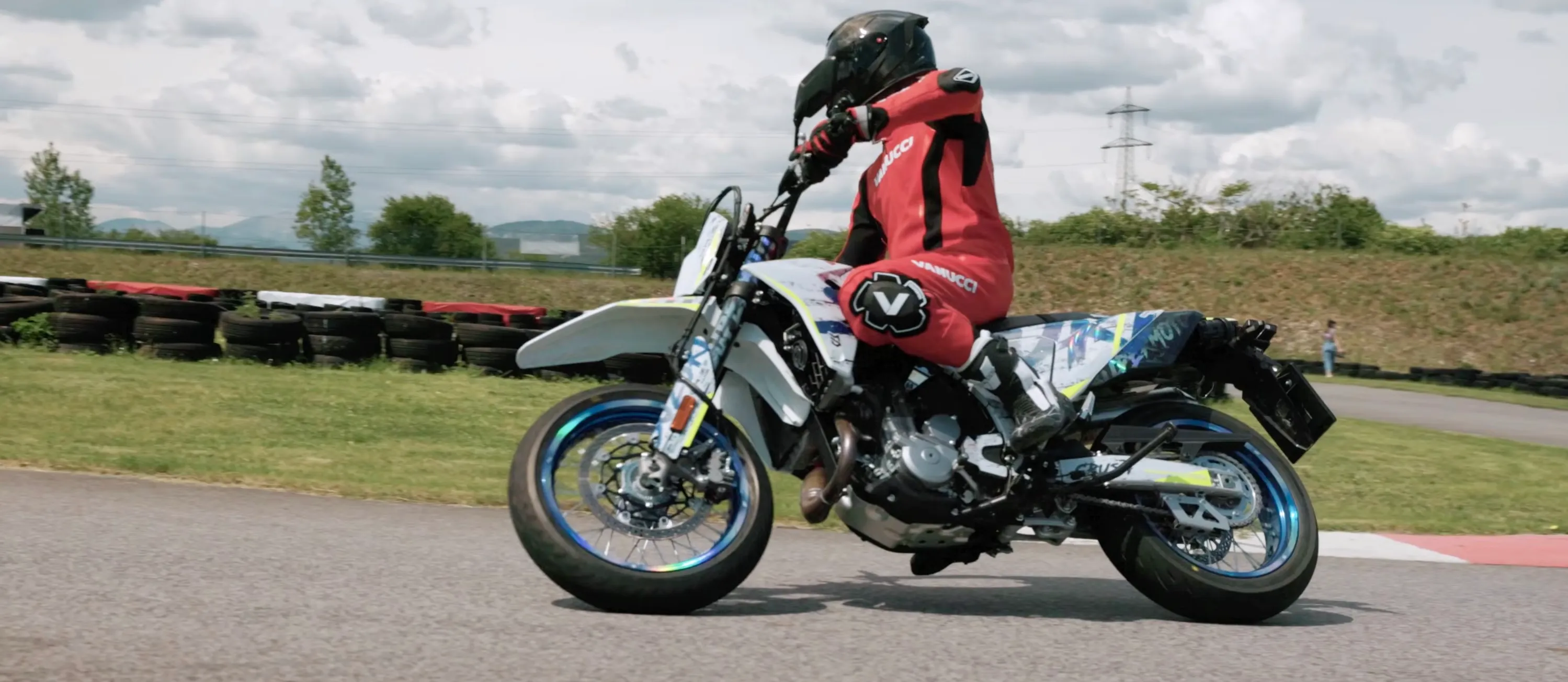 NastyNils rides the Suzuki DR-Z4SM with blue and white graphics through a corner at the Bad Fischau supermoto circuit in Austria, wearing red Vanucci leathers and a black helmet. No other motorcycle is visible.