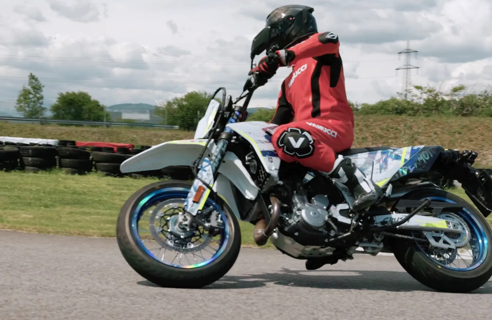 NastyNils rides the Suzuki DR-Z4SM with blue and white graphics through a corner at the Bad Fischau supermoto circuit in Austria, wearing red Vanucci leathers and a black helmet. No other motorcycle is visible.