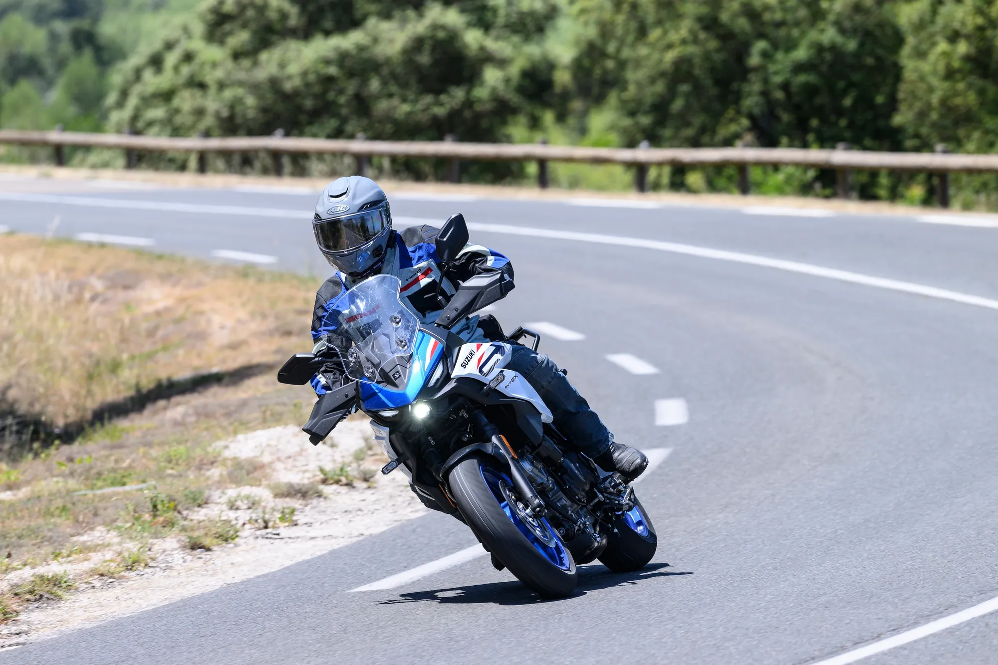 NastyNils rides the 2026 Suzuki SV-7GX through a fast right-hand sweeper on a tree-lined country road near Montpellier, headlight on, mid-corner lean.